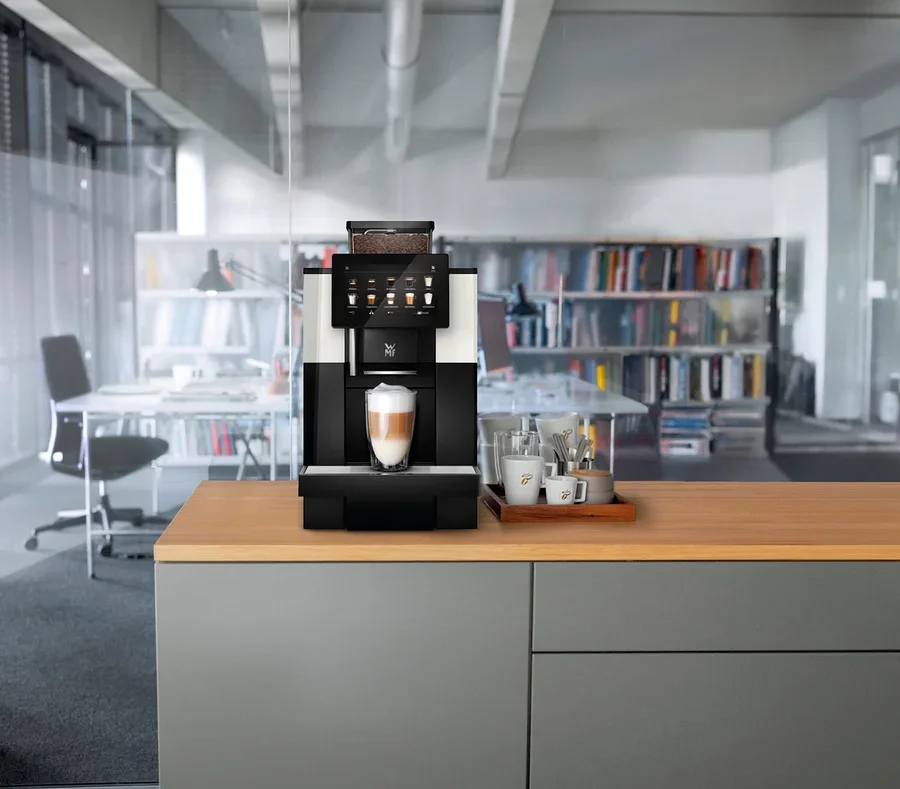 Ein Kaffeevollautomat steht auf einer Küchentheke mit einer Tasse Latte Macchiato davor. Daneben stehen Tassen und Gläser auf einem Holztablett.