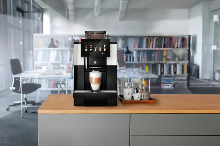 Ein Kaffeevollautomat steht auf einer Küchentheke mit einer Tasse Latte Macchiato davor. Daneben stehen Tassen und Gläser auf einem Holztablett.