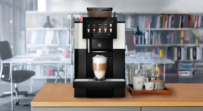 Ein Kaffeevollautomat steht auf einer Küchentheke mit einer Tasse Latte Macchiato davor. Daneben stehen Tassen und Gläser auf einem Holztablett.
