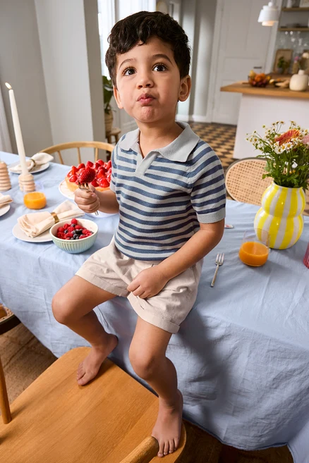 Junge sitzt auf Tischkante mit Erdbeere am Spieß, trägt Kinder-Shorts und Kinder-Strick-Poloshirt. Auf dem Tisch stehen Vase und Tortenplatte.