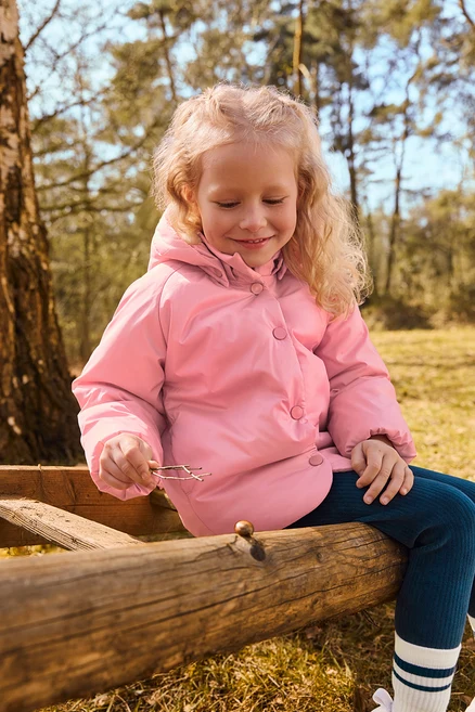 Ein kleines Mädchen mit blonden Locken, das eine rosa Steppjacke und dunkelblaue Leggings trägt, sitzt auf einem Holzzaun im Wald, hält kleine Äste und blickt auf den Zaun.