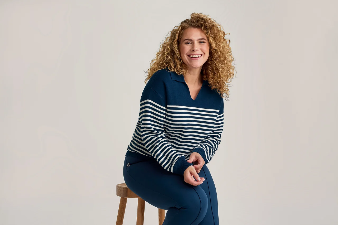 Lachende Frau mit Locken, die einen marineblauen und weiß gestreiften Pullover und marineblaue Hosen trägt, sitzt auf einem Holzschemel.
