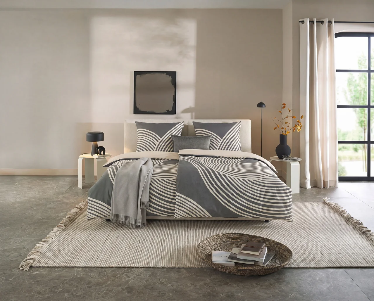 Modernes Schlafzimmer mit einem Bett, das mit der SCHÖNER WOHNEN – Kollektion Bettwäsche in Grau mit weißem Wellenmuster bezogen ist. Helle Nachttische, ein Teppich und ein Korb mit Büchern ergänzen den Raum.