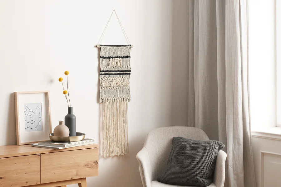 Ein Makramee-Wandteppich hängt an der Wand. Darunter steht eine Kommode, auf der eine Vase mit Blumen und ein Zierrahmen liegen.