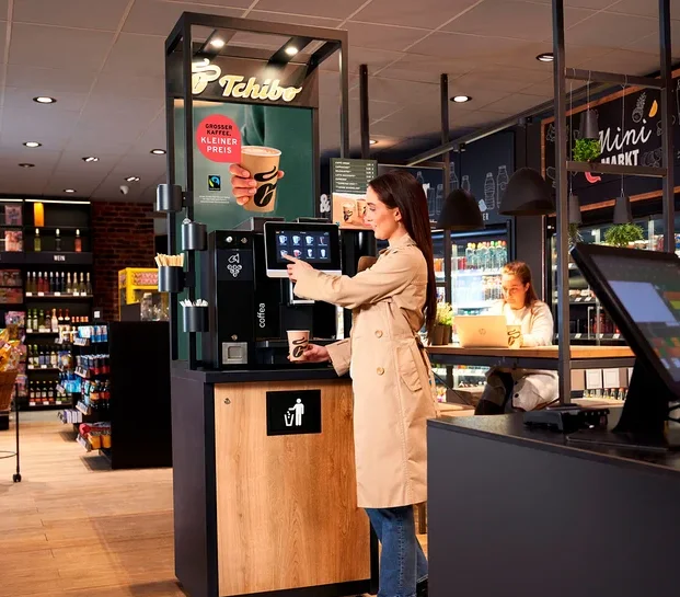 Eine Frau im beigen Trenchcoat bedient eine Tchibo Selbstbedienungs-Kaffeemaschine und hält einen Becher. Im Hintergrund arbeitet eine andere Frau an einem Laptop an einem Tresen im Minimarkt.