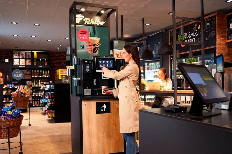 Eine Frau im beigen Trenchcoat bedient eine Tchibo Selbstbedienungs-Kaffeemaschine und hält einen Becher. Im Hintergrund arbeitet eine andere Frau an einem Laptop an einem Tresen im Minimarkt.
