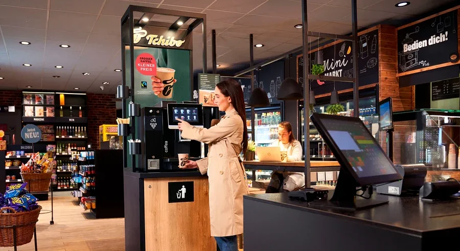 Eine Frau im beigen Trenchcoat bedient eine Tchibo Selbstbedienungs-Kaffeemaschine und hält einen Becher. Im Hintergrund arbeitet eine andere Frau an einem Laptop an einem Tresen im Minimarkt.