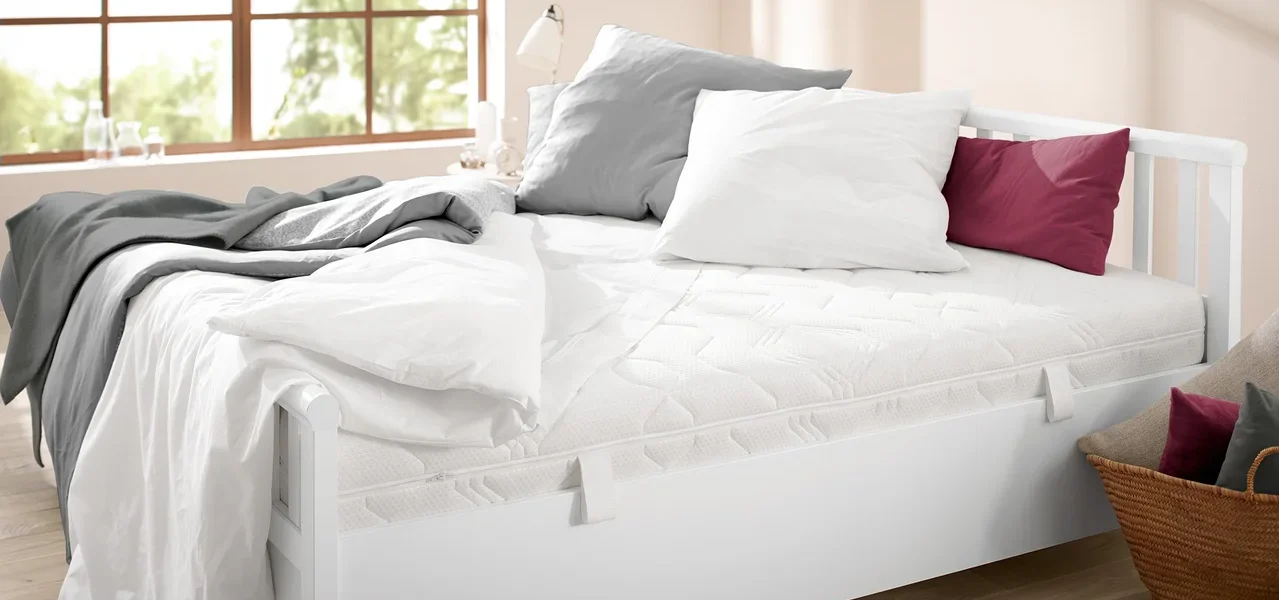 Bett mit weißem Rahmen und gesteppter Matratze. Auf dem Bett liegen eine graue Decke, ein weißes und ein graues Kissen sowie ein weinrotes Kissen.