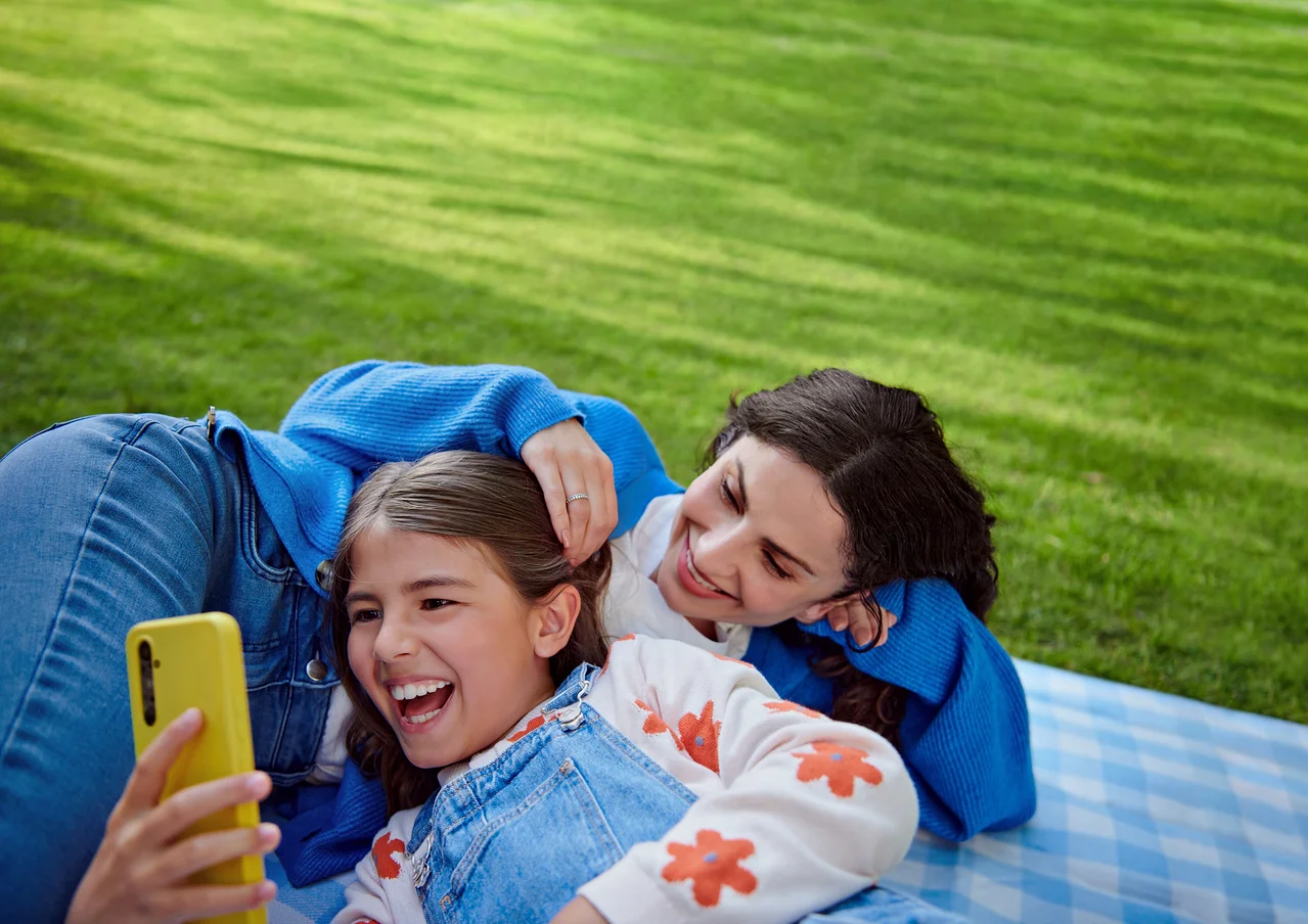 Mutter und Tochter liegen auf einer Decke im Gras. Das Mädchen lacht und hält ein gelbes Telefon.