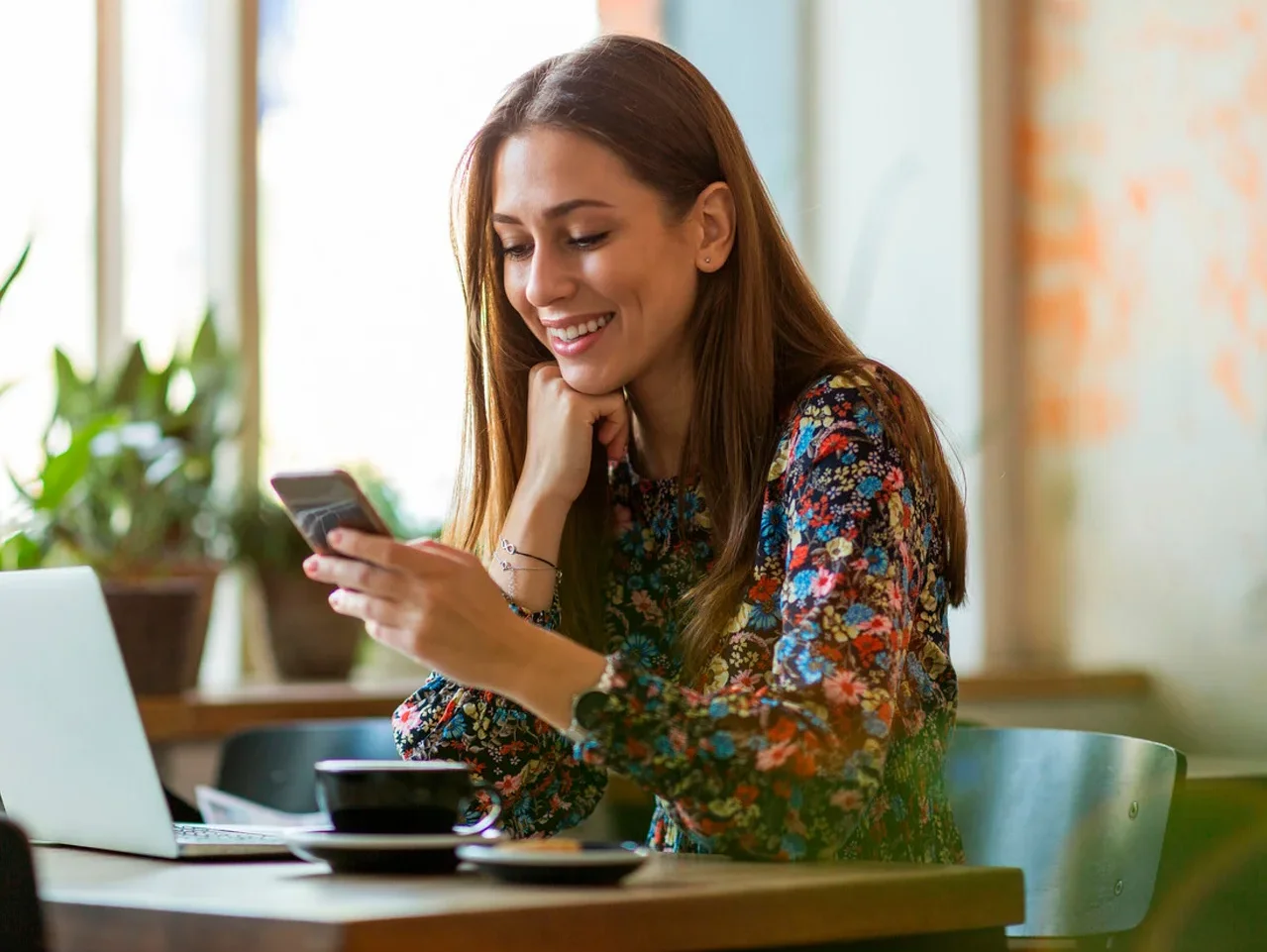 Frau lächelt und schaut auf ihr Mobiltelefon, sitzt an einem Tisch mit Laptop und einer Tasse Kaffee.