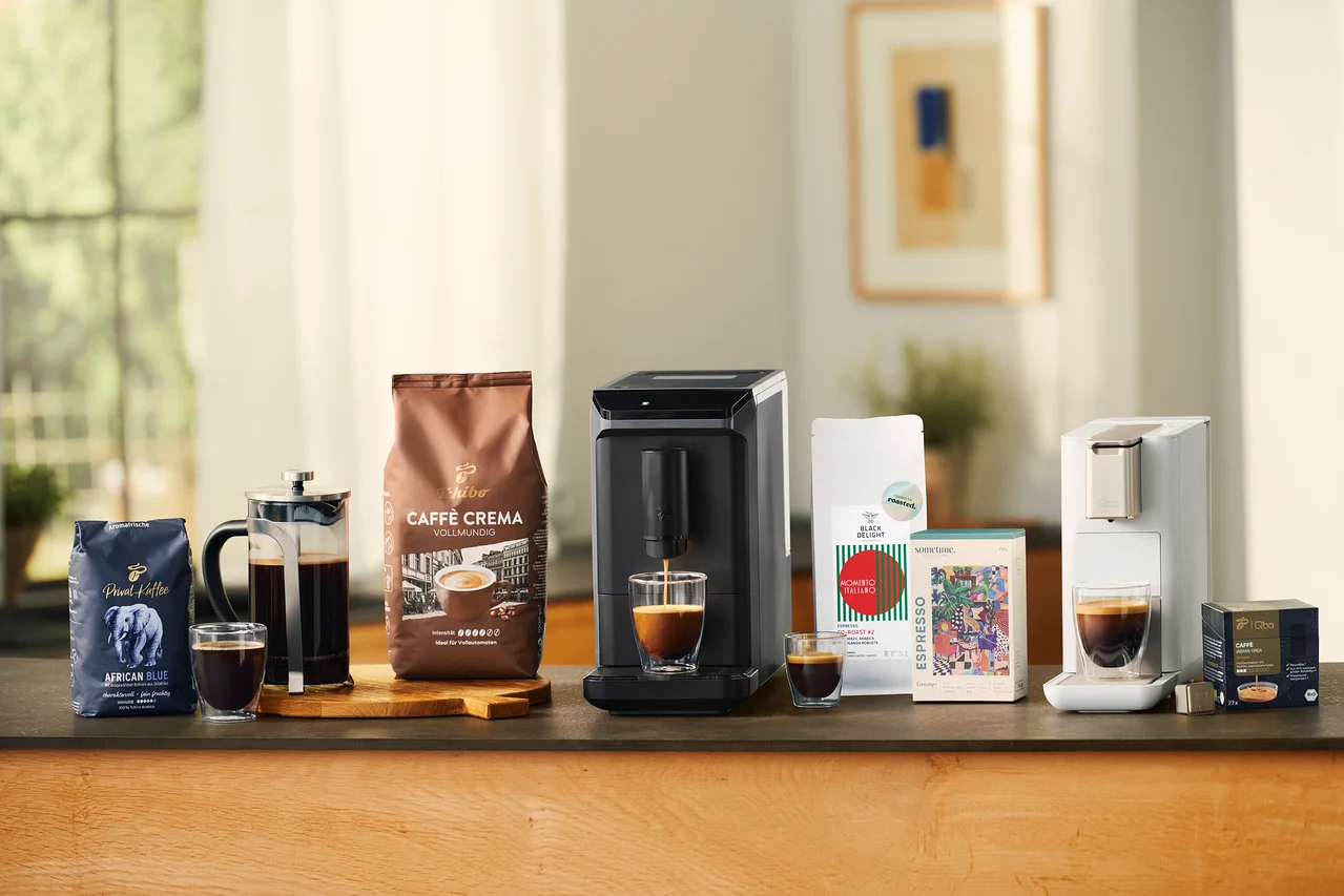 Verschiedene Kaffeeprodukte auf einer Theke: eine blaue Packung Privat Kaffee African Blue, eine French Press mit Kaffee, ein doppelwandiges Caffè Crema Glas, eine braune Packung Caffè Crema Vollmundig, ein grauer Esperto mini Kaffeevollautomat, der Kaffee in ein doppelwandiges Caffè Crema Glas füllt, ein kleines doppelwandiges Caffè Crema Glas, eine weiße Packung Black Delight Espresso, eine bunte sometime CONTEMP 1 Espresso Packung, eine weiße Qbo ICONIC Light Gold Maschine, die Kaffee in ein doppelwandiges Caffè Crema Glas füllt, und eine schwarze Packung Qbo Caffè JABANA YIRGA Kapseln.