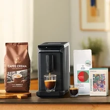 Verschiedene Kaffeeprodukte auf einer Theke: eine blaue Packung Privat Kaffee African Blue, eine French Press mit Kaffee, ein doppelwandiges Caffè Crema Glas, eine braune Packung Caffè Crema Vollmundig, ein grauer Esperto mini Kaffeevollautomat, der Kaffee in ein doppelwandiges Caffè Crema Glas füllt, ein kleines doppelwandiges Caffè Crema Glas, eine weiße Packung Black Delight Espresso, eine bunte sometime CONTEMP 1 Espresso Packung, eine weiße Qbo ICONIC Light Gold Maschine, die Kaffee in ein doppelwandiges Caffè Crema Glas füllt, und eine schwarze Packung Qbo Caffè JABANA YIRGA Kapseln.