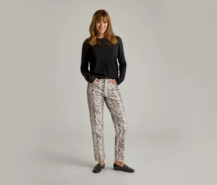 Frau steht lächelnd mit Händen in den Taschen, trägt ein schwarzes Langarmshirt, helle Hose mit Schlangenmuster und schwarze Loafer.