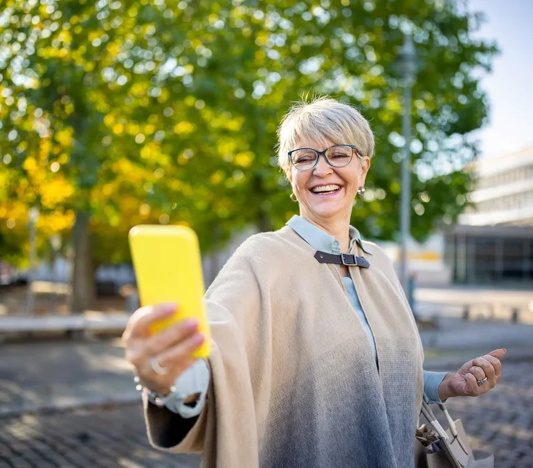 Frau mit Brille und beigem Poncho macht ein Selfie mit gelbem Handy im Freien.