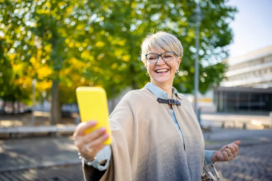Frau mit Brille und beigem Poncho macht ein Selfie mit gelbem Handy im Freien.