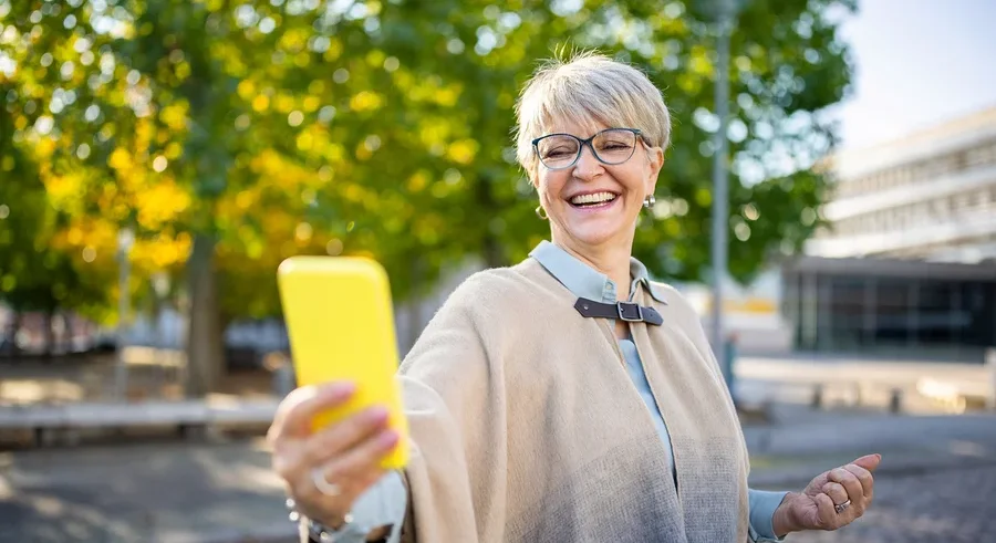 Frau mit Brille und beigem Poncho macht ein Selfie mit gelbem Handy im Freien.