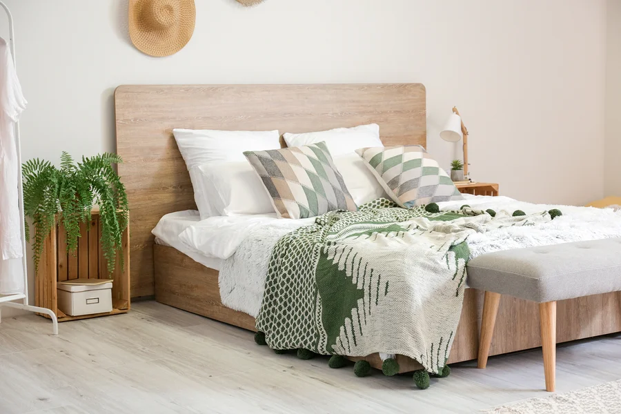 Bett mit weißer Bettwäsche, geometrischen Kissen und einer grün-weißen Decke mit Bommeln. Ein Holzhocker steht am Fußende.