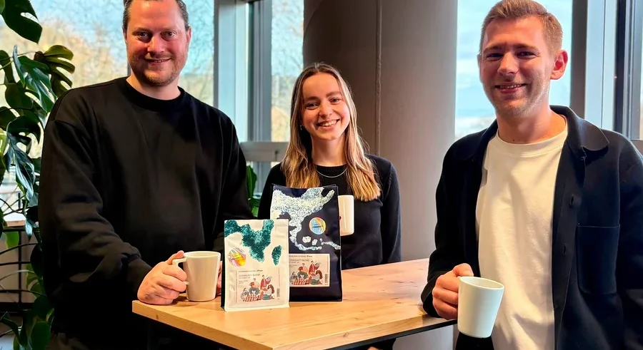 Drei Personen stehen an einem Tisch und halten Tassen. Auf dem Tisch stehen zwei Packungen Kaffee.