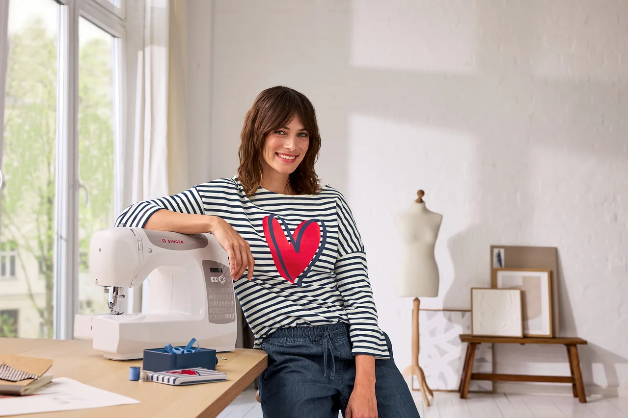 Eine lächelnde Frau in einem gestreiften Shirt mit rotem Herz lehnt an einer weißen Singer Nähmaschine auf einem Holztisch. Links ist ein Fenster, rechts eine Schaufensterpuppe und Bilder.