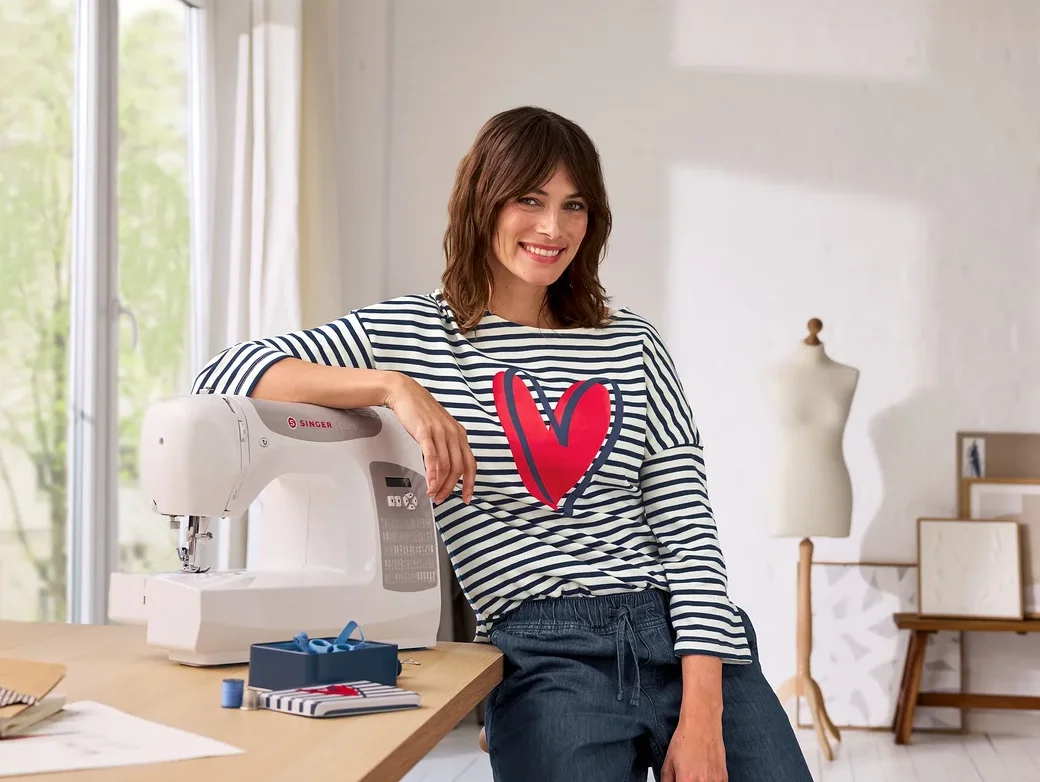Eine lächelnde Frau in einem gestreiften Shirt mit rotem Herz lehnt an einer weißen Singer Nähmaschine auf einem Holztisch. Links ist ein Fenster, rechts eine Schaufensterpuppe und Bilder.