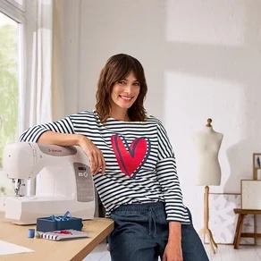Eine lächelnde Frau in einem gestreiften Shirt mit rotem Herz lehnt an einer weißen Singer Nähmaschine auf einem Holztisch. Links ist ein Fenster, rechts eine Schaufensterpuppe und Bilder.