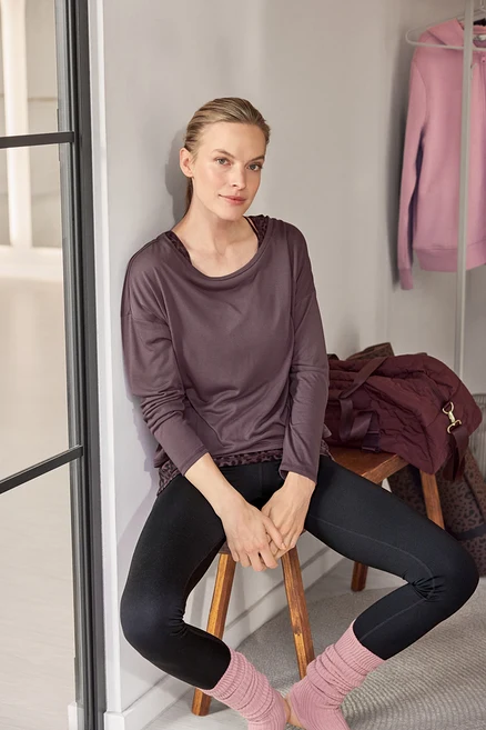 Eine Frau sitzt auf einem Hocker, lehnt an einer Wand und trägt ein dunkles Langarmshirt mit Leopardenmuster, schwarze Leggings und rosa Stulpen.