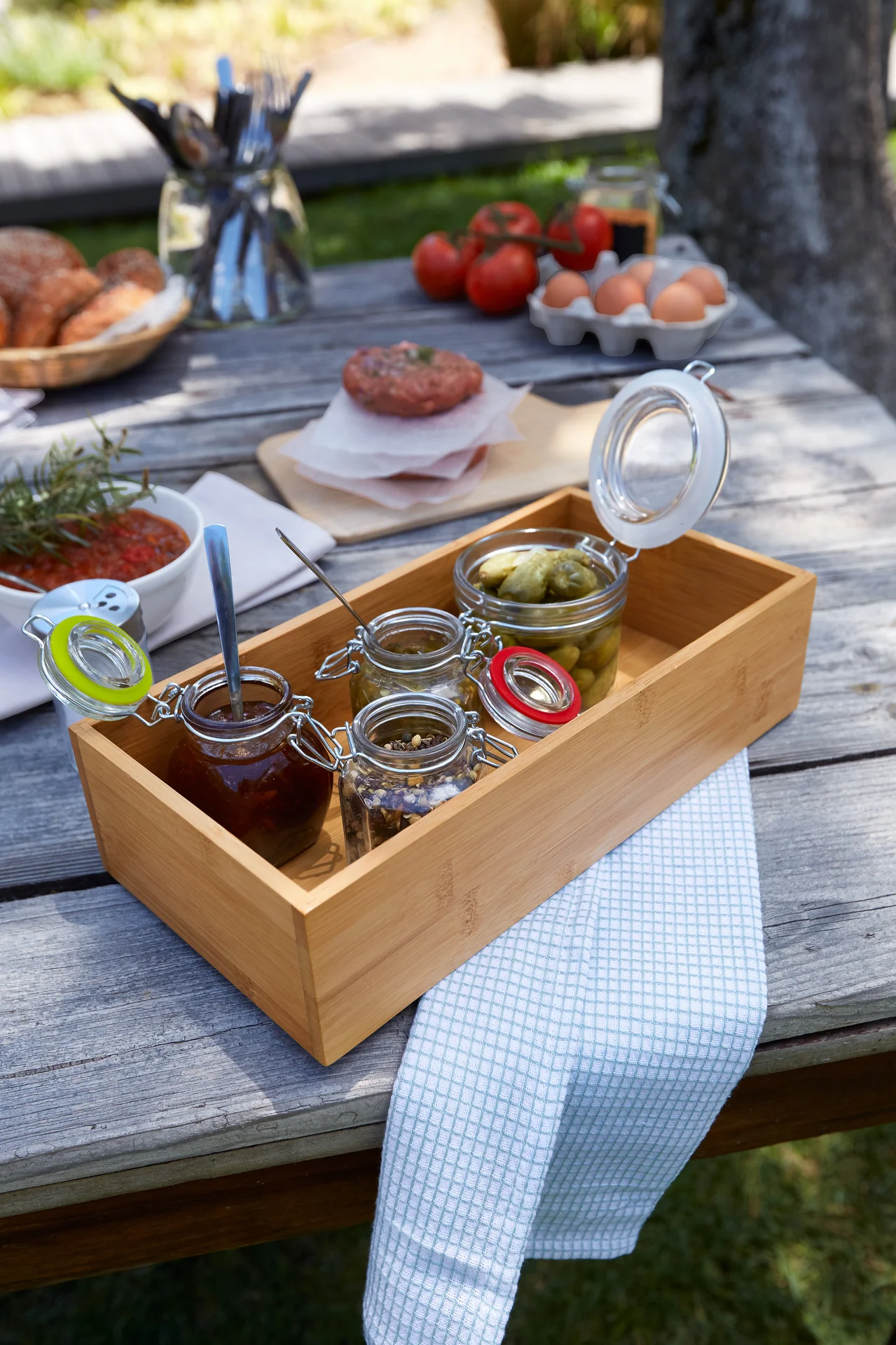 Auf einem Holztisch steht ein Tablett mit kleinen Gläsern mit verschiedenen Lebensmitteln, daneben eine Schüssel mit Soße, Fleisch, Eier und Brot.