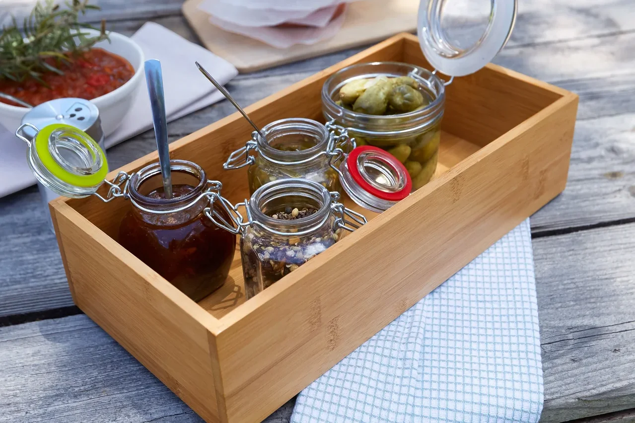 Auf einem Holztisch steht ein Tablett mit kleinen Gläsern mit verschiedenen Lebensmitteln, daneben eine Schüssel mit Soße, Fleisch, Eier und Brot.