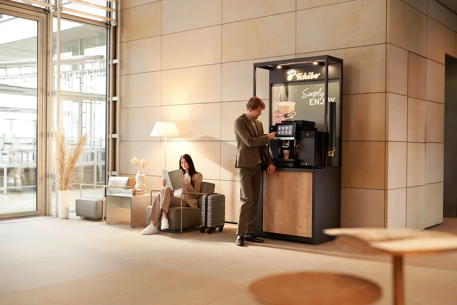 Ein Mann bedient eine Kaffeemaschine, während eine Frau mit einem Laptop in einem Sessel neben einem Koffer sitzt.