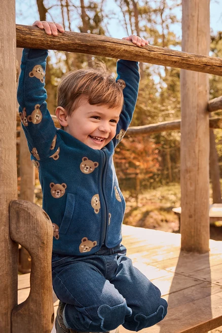 Ein lächelnder kleiner Junge hängt an einer Holzstange, trägt eine blaue Fleecejacke mit Bärenmustern und blaue Jeans mit bärenförmigen Knieapplikationen, auf einem Spielplatz im Freien.