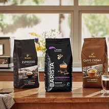 Auf einem Holztisch stehen drei Tchibo Kaffeepackungen nebeneinander: Espresso Kräftig, Barista Espresso Dark und Caffè Crema Vollmundig. Im Hintergrund befinden sich eine Kaffeemaschine und ein Fenster.