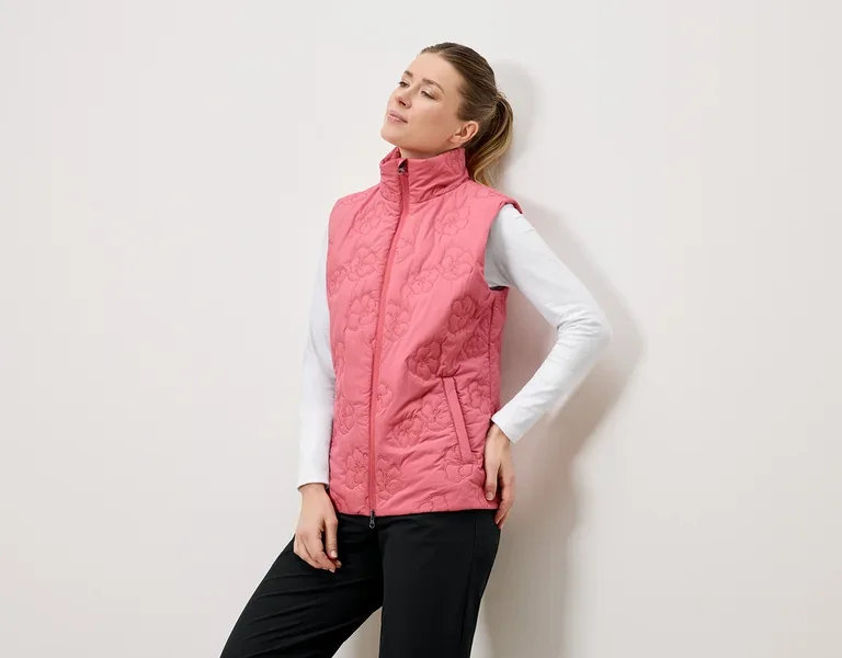 Eine Frau posiert in einer gesteppten, rosafarbenen Weste mit Blumenmuster, einem weißen Langarmshirt und schwarzen Hosen vor einer weißen Wand.