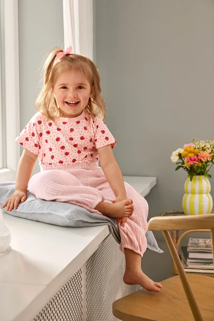 Ein lächelndes Mädchen sitzt in einem 2 Kinder-T-Shirts und einer Kinder-Musselin-Hose auf einem Kissen. Eine Vase steht auf einem Stuhl.