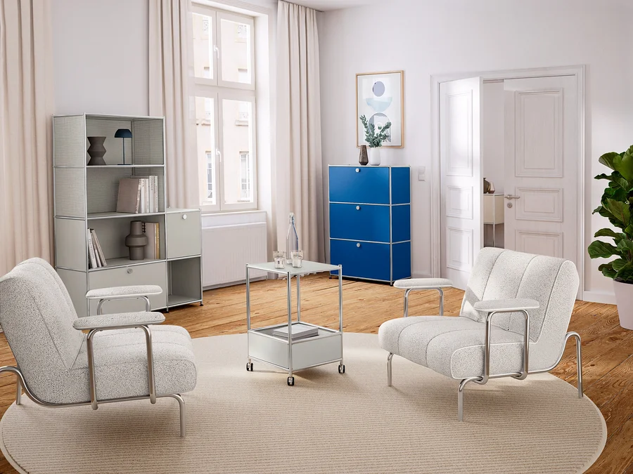 Helles Wohnzimmer mit Holzboden und weißer Einrichtung. Links ein hellgraues Regal, in der Mitte zwei helle Sessel und ein Beistelltisch auf einem runden Teppich. Rechts ein blauer Schrank und eine Pflanze.