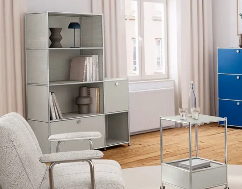 Helles Wohnzimmer mit Holzboden und weißer Einrichtung. Links ein hellgraues Regal, in der Mitte zwei helle Sessel und ein Beistelltisch auf einem runden Teppich. Rechts ein blauer Schrank und eine Pflanze.