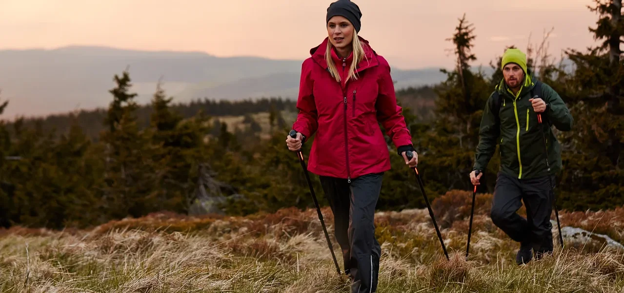 Zwei Personen wandern mit Stöcken über eine Wiese.