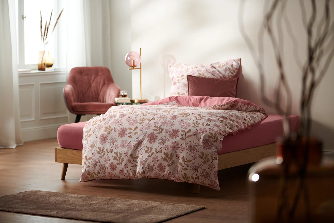Blumige Bettwäsche auf einem Bett mit rosa Spannbettlaken. Darunter Renforcé-Bettwäsche in Normal- und Übergröße.