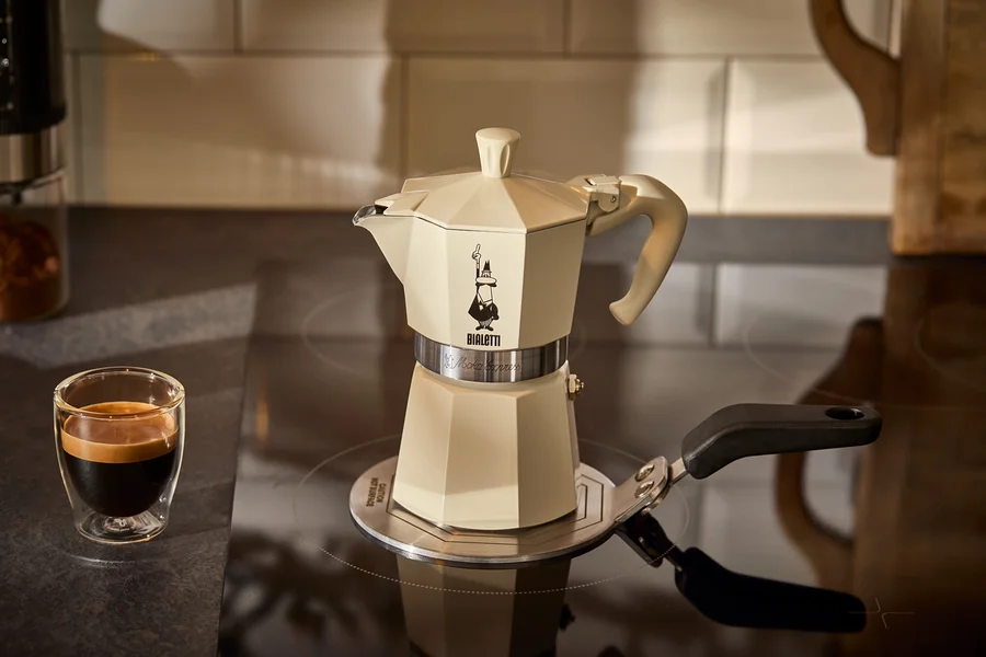 Ein cremefarbener Espressokocher Bialetti Moka Express 190 ml steht auf einer Bialetti Adapterplatte Induktion 13 cm neben einem Glas Espresso.