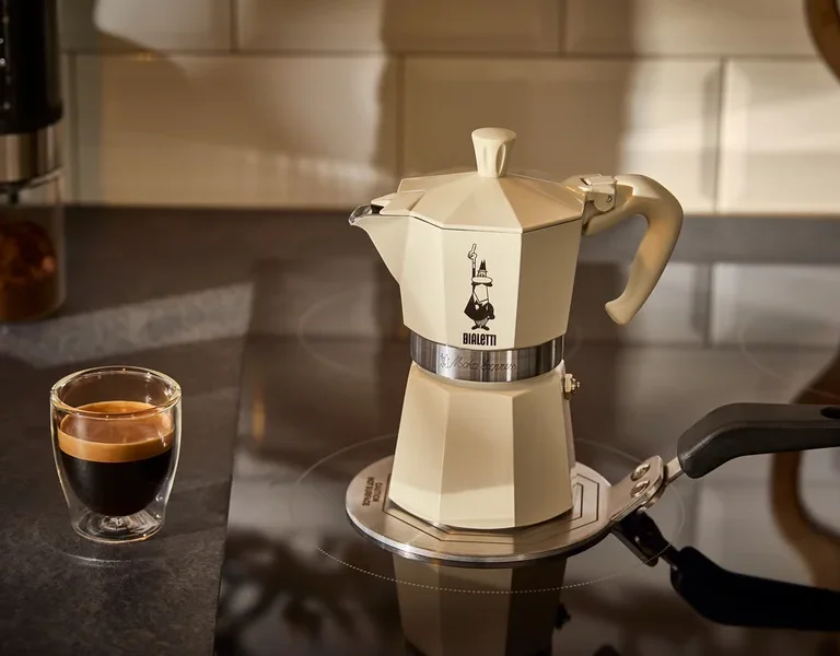 Ein cremefarbener Espressokocher Bialetti Moka Express 190 ml steht auf einer Bialetti Adapterplatte Induktion 13 cm neben einem Glas Espresso.