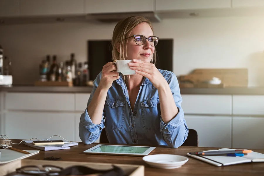 Frau mit Brille sitzt an einem Holztisch und hält eine Tasse in der Hand. Auf dem Tisch liegen ein Tablet, ein Notizbuch und Stifte.