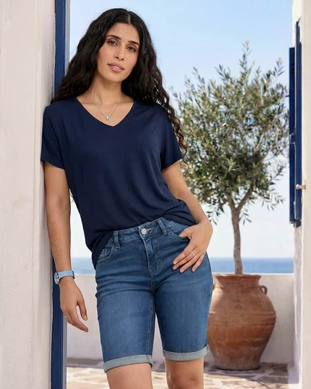 Frau lehnt an Wand, trägt dunkelblaues Shirt, Bermuda-Jeans, blaue und transparente Ringe, Armbanduhr mit Lederband.