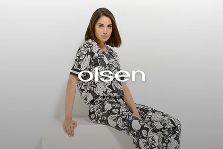 Eine Frau mit braunen Haaren sitzt in einem schwarz-weißen, floral gemusterten Olsen-Outfit vor einem weißen Hintergrund.