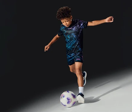 Ein Junge in einem hummel® »HML Blaze Graphic Set Kids« spielt Fußball und tritt einen weiß-lila Ball.