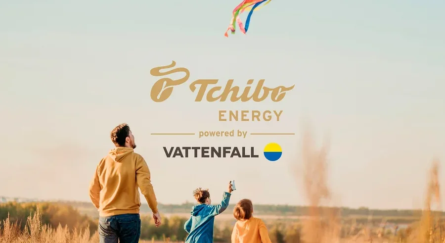Ein Mann und zwei Kinder laufen durch ein Feld mit hohem, trockenem Gras und blicken zu einem bunten Drachen am klaren Himmel auf. Der Mann trägt einen gelben Hoodie und Jeans. Ein Kind trägt eine blaue Jacke, das andere ein gelbes Oberteil. Ein "Tchibo ENERGY powered by VATTENFALL" Logo ist am Himmel sichtbar.