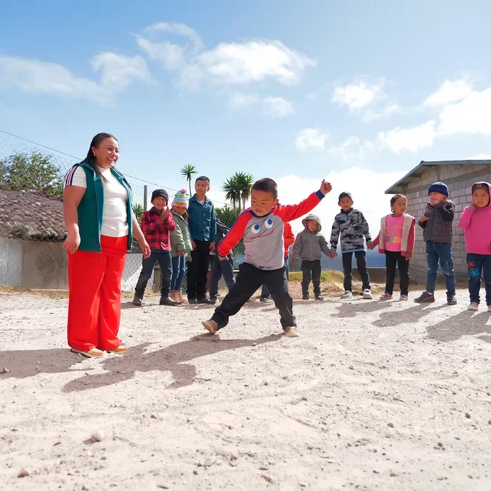 Eine Frau in grüner Weste und roter Hose lächelt einen Jungen in grau-rotem Shirt an, der auf einem Sandplatz tritt. Mehrere Kinder beobachten sie im Hintergrund unter blauem Himmel.