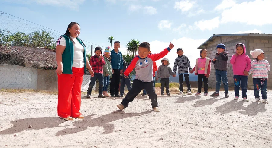 Eine Frau in grüner Weste und roter Hose lächelt einen Jungen in grau-rotem Shirt an, der auf einem Sandplatz tritt. Mehrere Kinder beobachten sie im Hintergrund unter blauem Himmel.