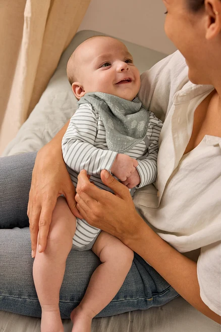Eine Mutter hält ein lächelndes Baby, das einen Baby-Body mit Mitwachsfunktion und ein Baby-Musselin-Halstuch trägt.