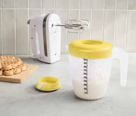 Ein weißer Mixer, ein Messbecher mit gelbem Deckel voller Flüssigkeit und ein Stapel Waffeln auf einem Brett stehen auf der Arbeitsfläche.