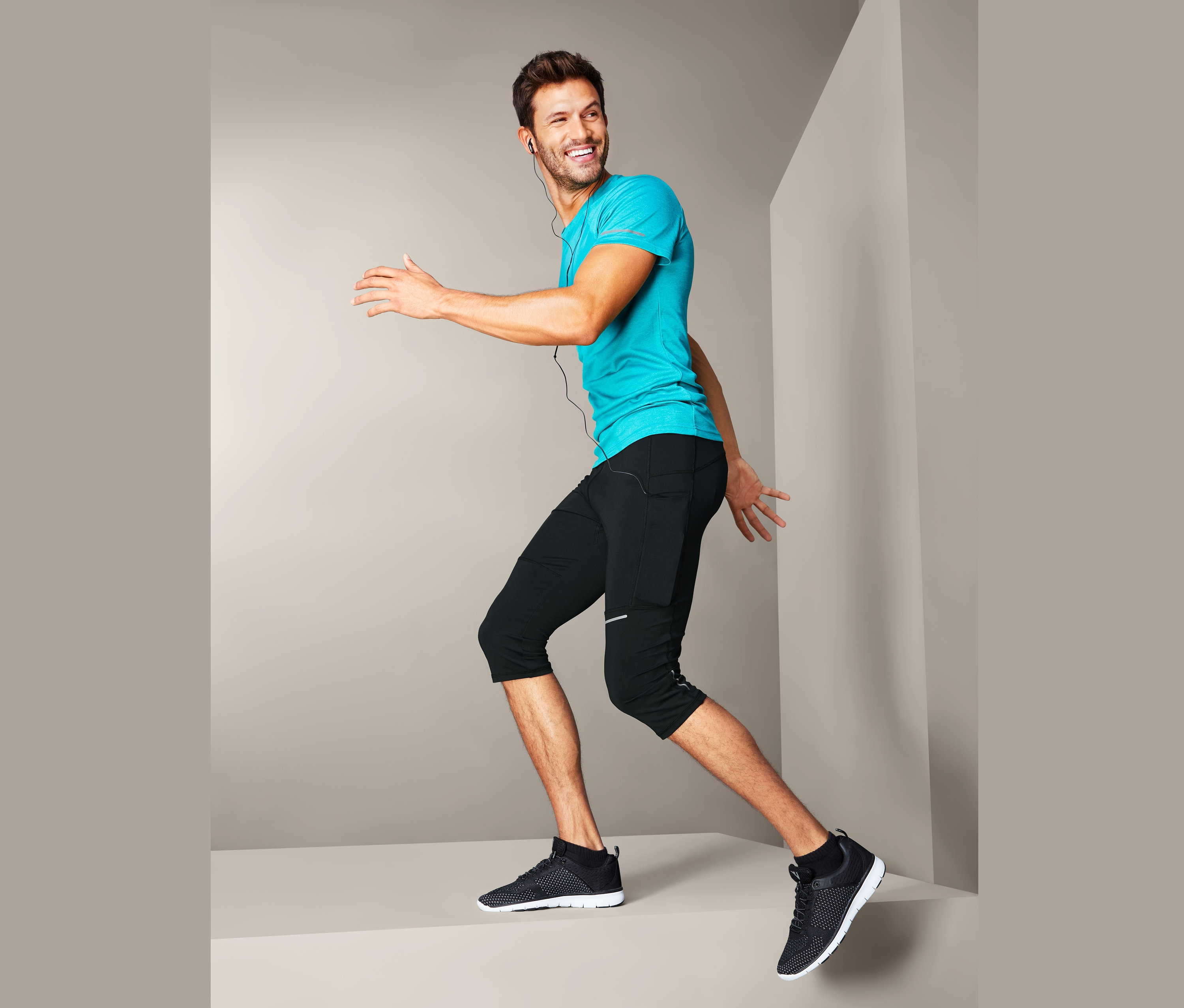 Lächelnder Mann in türkisfarbenem T-Shirt und 3/4-Lauftight posiert vor grauem Hintergrund.