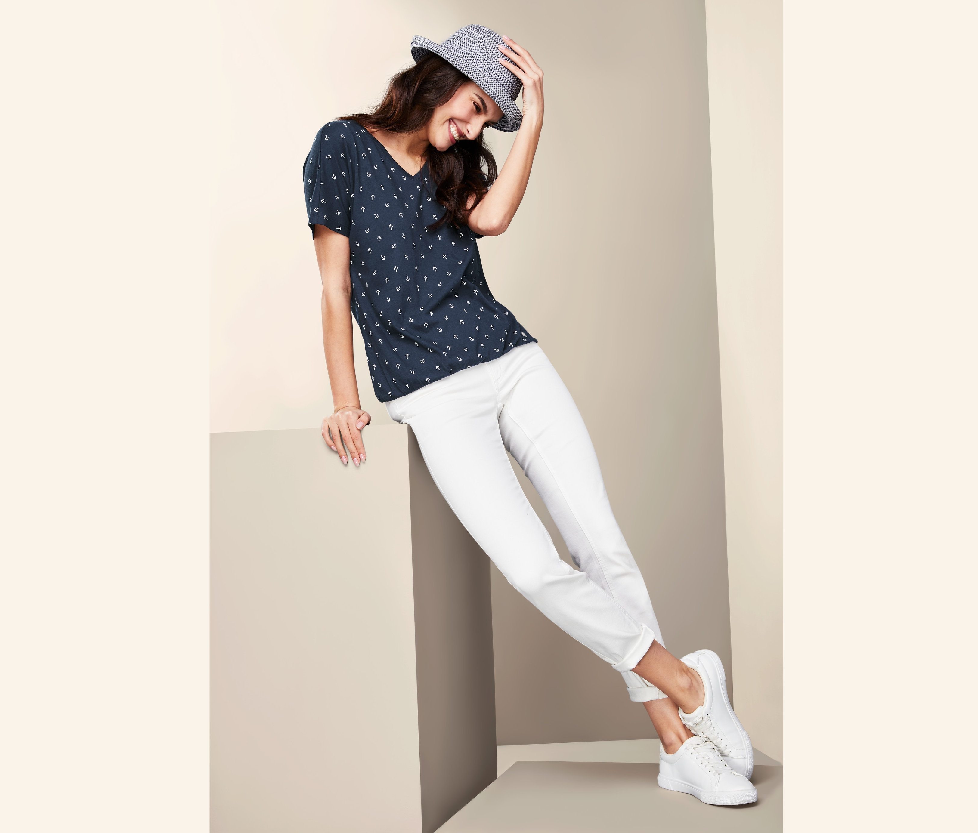 Frau lächelt und hält einen Hut auf dem Kopf, trägt ein Blusenshirt und eine weisse Hose.